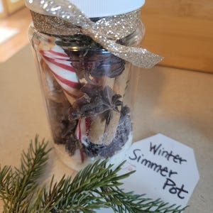 May include: A clear jar filled with winter simmer pot ingredients, including cinnamon sticks, star anise, and candy canes. The jar is topped with a white lid and a gold glitter ribbon. A tag reads "Winter Simmer Pot."