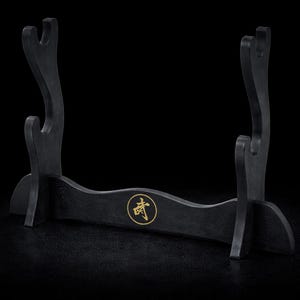 May include: A black wooden sword stand with a curved design. The stand features two upright supports with curved tops and a central horizontal piece with a gold emblem. The stand is designed to hold a sword.