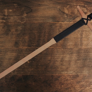 May include: A wooden sword with a black leather handle and a cross guard. The sword is lying on a wooden surface.