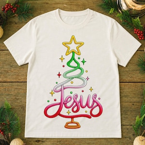 Op de afbeelding: Een crèmekleurig T-shirt met korte mouwen en een afbeelding van een gestileerde kerstboom gevormd door een doorlopende lijn. Het woord Jesus staat in rood cursief schrift aan de basis, bekroond met een gele ster en kleurrijke accenten.
