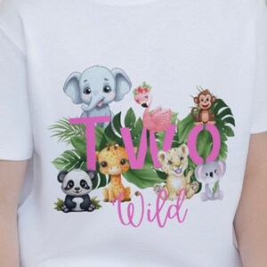 K&ouml;nnte beinhalten: Wei&szlig;es T-Shirt mit einer pinkfarbenen Grafik "TWO WILD", die verschiedene Dschungeltiere zeigt, darunter einen Elefanten, eine Giraffe, einen L&ouml;wen, einen Panda, einen Koala und einen Flamingo.