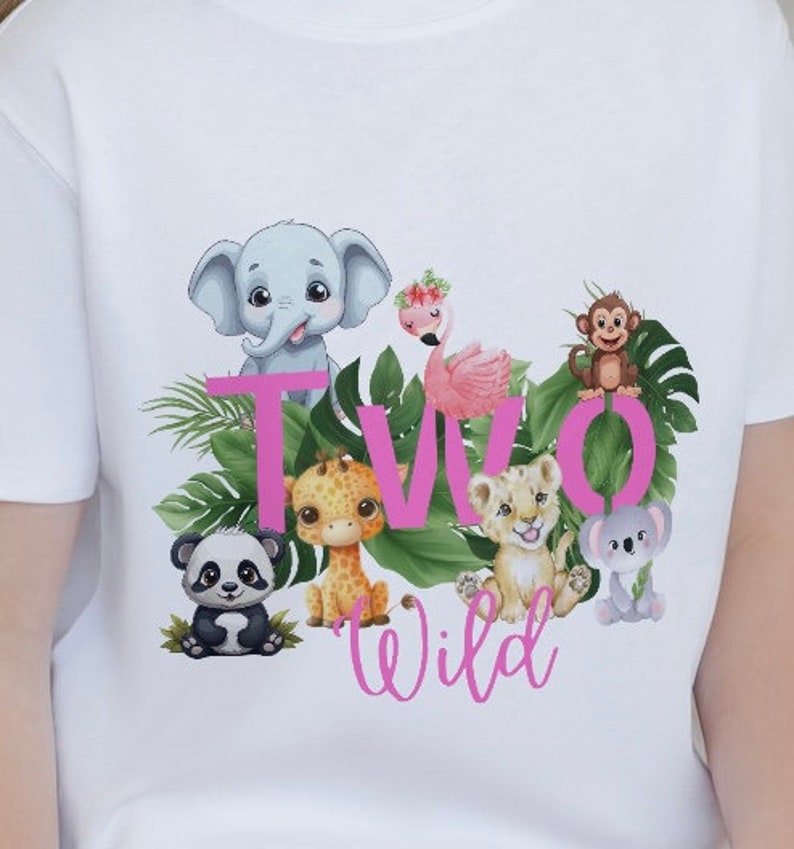K&ouml;nnte beinhalten: Wei&szlig;es T-Shirt mit einer pinkfarbenen Grafik "TWO WILD", die verschiedene Dschungeltiere zeigt, darunter einen Elefanten, eine Giraffe, einen L&ouml;wen, einen Panda, einen Koala und einen Flamingo.