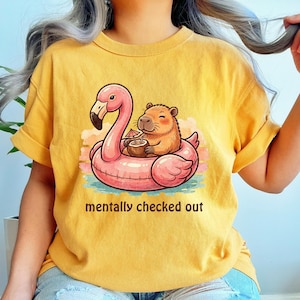 Könnte beinhalten: Senfgelbes T-Shirt mit einer Cartoon-Illustration eines Capybaras auf einem rosa Flamingo-Schwimmring, der einen Cocktail hält. Der Text "mentally checked out" ist darunter gedruckt. Tropisches Thema.