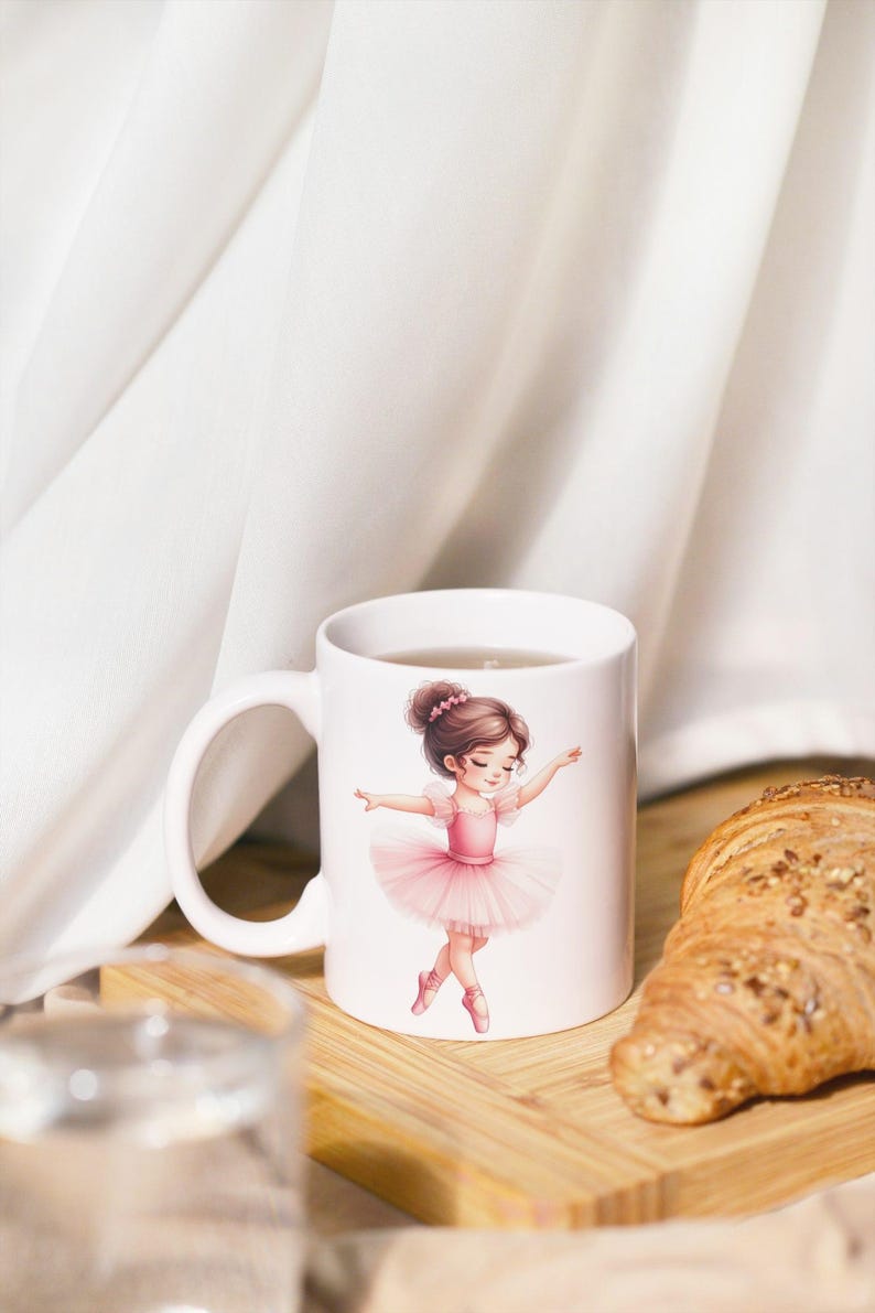 Op de afbeelding: Witte keramische mok met een ballerina in een roze tutu. De illustratie toont een jong meisje in een balletpose. De mok staat op een houten plank met een croissant. De achtergrond is een wit gordijn.