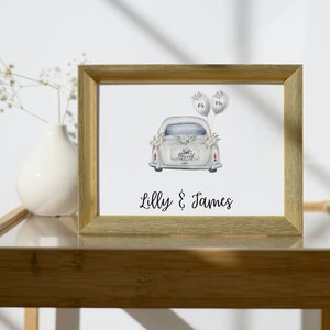 May include: A gold-framed print with a watercolor illustration of a white car with "Just Married" on the back. The car has two white balloons tied to the back. The print has the text "Lilly & James" at the bottom.