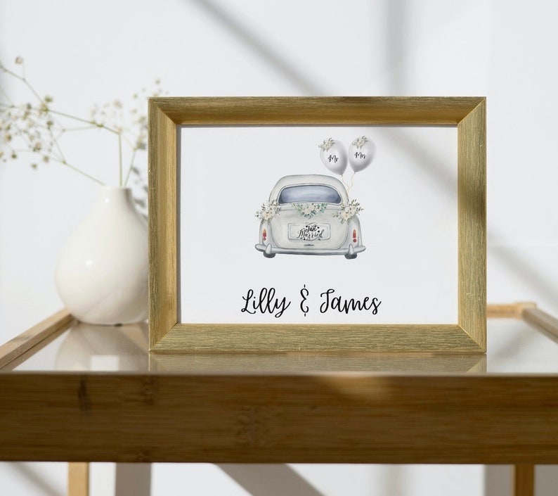 May include: A gold-framed print with a watercolor illustration of a white car with "Just Married" on the back. The car has two white balloons tied to the back. The print has the text "Lilly & James" at the bottom.