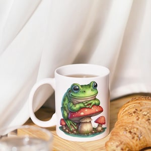 May include: White ceramic mug featuring a green frog sitting on a red mushroom with white spots. The frog has large, expressive eyes. The mug is on a wooden surface, with a croissant and a glass of water in the background.