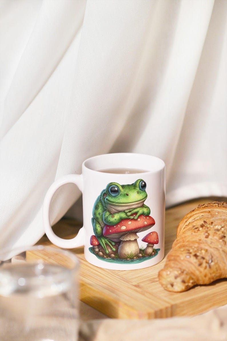 May include: White ceramic mug featuring a green frog sitting on a red mushroom with white spots. The frog has large, expressive eyes. The mug is on a wooden surface, with a croissant and a glass of water in the background.