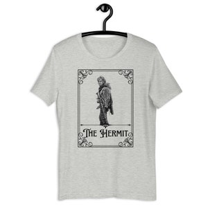 May include: A heather grey t-shirt with a black and white illustration of a person holding a rifle. The illustration is framed with a decorative border and the text "The Hermit" is printed below the image.