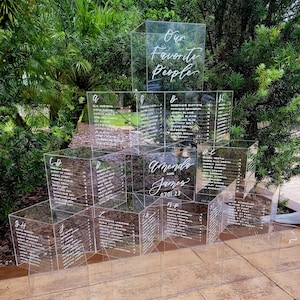 May include: Clear acrylic wedding seating chart with multiple cube-shaped sections. The chart features names and table assignments in white script. The top section reads "Our Favorite People". The names "Amanda and James" and the date "07.01.23" are also displayed.