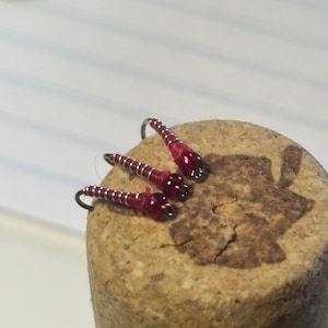 Può includere: Tre esche da pesca rosse e argentate con ami neri poggiano su un tappo di sughero. Le esche hanno un design segmentato con un corpo rosso lucido e dettagli argentati. Il tappo di sughero ha un aspetto naturale e strutturato.