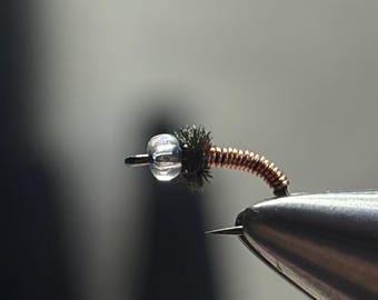 May include: A fishing fly lure featuring a clear glass bead at the front, followed by a small dark brown fuzzy thorax section, and a curved body wrapped in thin copper wire. The lure is attached to a metal hook, showing intricate construction details.