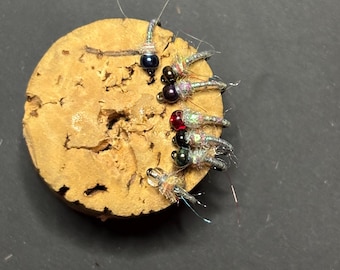 Könnte beinhalten: Ein Korkbrett mit sechs Fliegenfischerködern mit unterschiedlich farbigen Perlen. Die Köder sind silberfarben mit schillernden Flügeln und schwarzen, roten und violetten Perlen.