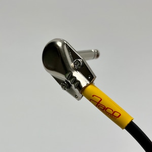 May include: Close-up of a silver right-angle guitar cable connector with a yellow cable and red text that reads "Jaco" on the cable.
