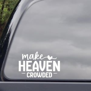 May include: White vinyl decal with the phrase "make Heaven CROWDED" in a script and block font. The decal is applied to a car window, with a blurred background of trees and sky.