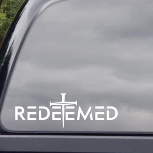 May include: White vinyl decal with the word "REDEEMED" and a cross design. The decal is applied to a vehicle window, with a cloudy sky visible in the background. The design is simple and bold.