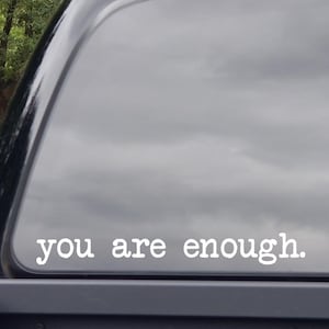 May include: White vinyl decal with the words "you are enough" in a simple, sans-serif font. The decal is affixed to the rear window of a black vehicle, with a cloudy sky and trees visible in the background.