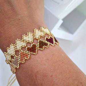Puede incluir: Una pulsera de cuentas con un diseño en forma de corazón. La pulsera presenta un borde dorado y está hecha con pequeñas cuentas en tonos crema, marrón y burdeos. La pulsera está atada con un cordón dorado.
