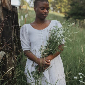 May include: A person wearing a white dress stands in a field of tall grass, holding a bouquet of white flowers. The person is looking down at the flowers.