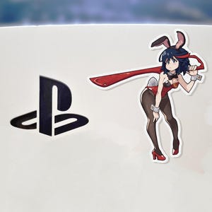 May include: White surface with a black PlayStation logo and a sticker of an anime character in a bunny outfit. The character is wearing a red outfit, black tights, and red heels. The sticker has a red ribbon.