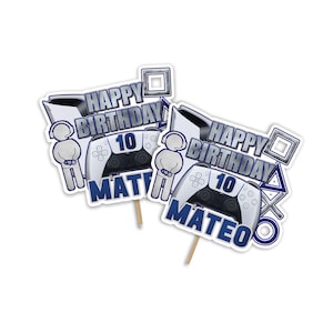 Puede incluir: Dos adornos para tartas blancos y azules con un mando de videojuegos, una figura que sostiene un mando y el texto "Feliz cumpleaños" y "Mateo" con el número "10".