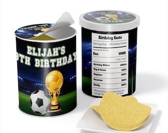 May include: A green, white, and black cylindrical container with a soccer ball and trophy graphic. The container has the text "Elijah's 8th Birthday" on the front. The container is open and has a label on the side that reads "Birthday Facts" with a list of fun facts about birthdays. The container is sitting on a white surface with a stack of yellow tortilla chips.