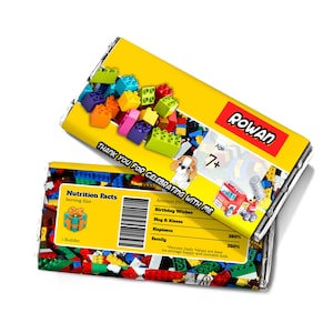Può includere: Un involucro di cioccolato giallo e rosso a tema Lego. L'involucro presenta un'etichetta di informazioni nutrizionali con il testo "Nutrition Facts" e "Serving Size: 1 Builder". L'involucro include anche il testo "Thank You For Celebrating With Me" e "Rowan Age 7+".
