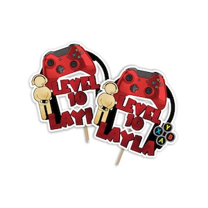 May include: Two red and black video game controller cake toppers with the text "LEVEL 10 LAYLA". Each topper features a red controller, a stick figure, and a black and white circular design.