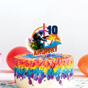 Può includere: Un topper per torta colorato con un giocatore di paintball, un bersaglio e il numero 10. Il testo "ANTHONY" è scritto sotto l'immagine.