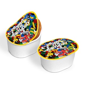 May include: Two white plastic containers with colorful labels. The labels feature a Lego theme with a red truck and the text "Rowan 7th Birthday".