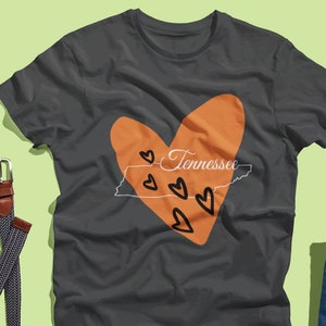 Op de afbeelding: Een donkergrijs T-shirt met een oranje hartvormig ontwerp met de omtrek van de staat Tennessee en het woord "Tennessee" in het hart. Het hart is omringd door kleinere oranje harten.