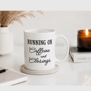May include: White ceramic mug with the text "RUNNING ON Caffeine and Closings" in black script. The mug sits on a round, light gray coaster. A lit candle and a vase with dried plants are in the background.
