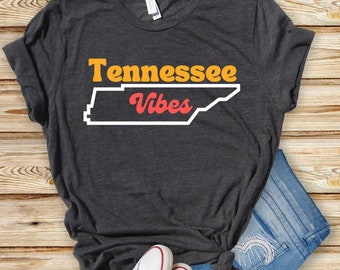 Könnte beinhalten: Ein dunkelgraues T-Shirt mit dem Text "Tennessee Vibes" in orangefarbenen und roten Buchstaben. Der Text befindet sich über einem Umriss des Bundesstaates Tennessee.