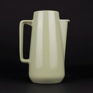 May include: A pale sage green ceramic pitcher with a smooth, tapered cylindrical body, a flat base, and a rectangular handle attached to the side. The pitcher features a simple, integrated spout at the top rim, maintaining a clean, uniform finish.