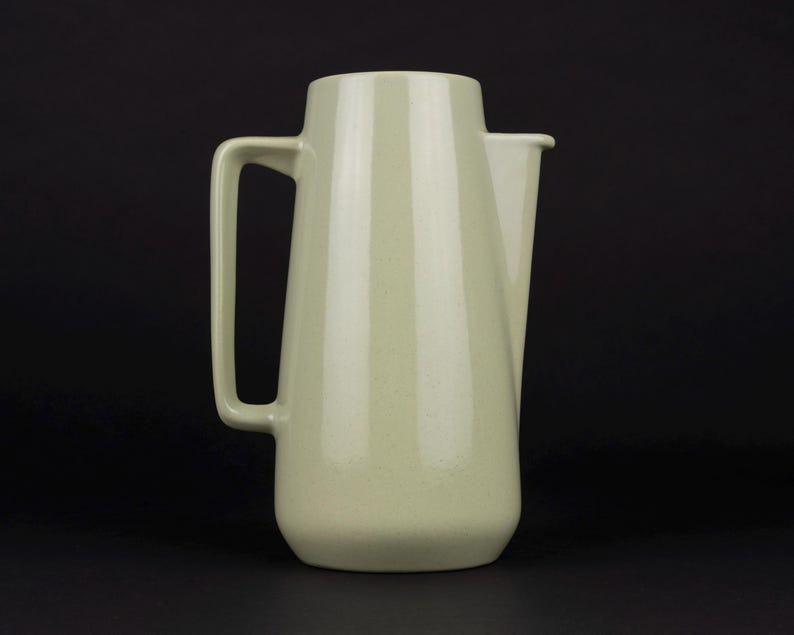 May include: A pale sage green ceramic pitcher with a smooth, tapered cylindrical body, a flat base, and a rectangular handle attached to the side. The pitcher features a simple, integrated spout at the top rim, maintaining a clean, uniform finish.