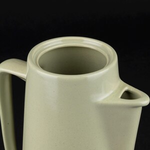 May include: A light sage green ceramic jug featuring a smooth, speckled finish, a wide circular opening at the top, a sturdy side handle, and a triangular-shaped pouring spout. The vessel has a clean, uniform design with a matte texture.