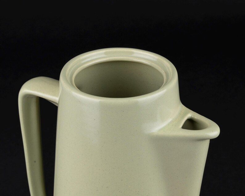 May include: A light sage green ceramic jug featuring a smooth, speckled finish, a wide circular opening at the top, a sturdy side handle, and a triangular-shaped pouring spout. The vessel has a clean, uniform design with a matte texture.
