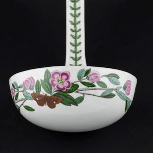 May include: A white ceramic ladle featuring a botanical illustration of pink flowers, green leaves, and a brown moth on the bowl. The handle is decorated with a repeating green leaf pattern. The design is colorful and detailed against a white background.