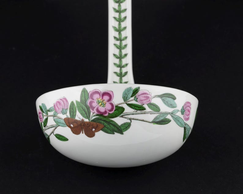 May include: A white ceramic ladle featuring a botanical illustration of pink flowers, green leaves, and a brown moth on the bowl. The handle is decorated with a repeating green leaf pattern. The design is colorful and detailed against a white background.