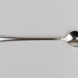 May include: A silver-toned metal spoon featuring an elongated handle with a decorative floral relief pattern at the tip. The spoon has a smooth, polished finish on the bowl and a slender neck that connects to the ornate handle end.