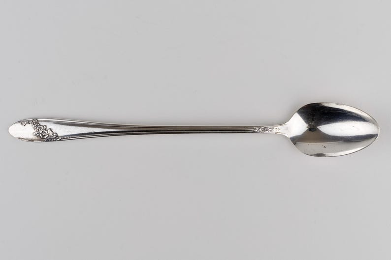May include: A silver-toned metal spoon featuring an elongated handle with a decorative floral relief pattern at the tip. The spoon has a smooth, polished finish on the bowl and a slender neck that connects to the ornate handle end.