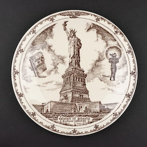 K&ouml;nnte beinhalten: Ein runder Keramikteller mit einer braunen Tuschezeichnung der Freiheitsstatue auf Bedloe's Island. Das Design enth&auml;lt Beschriftungen f&uuml;r Tablet und Torch sowie einen Rand mit Sternen und Adlern. Der Text lautet Statue of Liberty, Bedloe's Island, NY.
