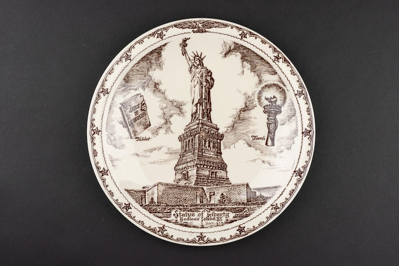 K&ouml;nnte beinhalten: Ein runder Keramikteller mit einer braunen Tuschezeichnung der Freiheitsstatue auf Bedloe's Island. Das Design enth&auml;lt Beschriftungen f&uuml;r Tablet und Torch sowie einen Rand mit Sternen und Adlern. Der Text lautet Statue of Liberty, Bedloe's Island, NY.
