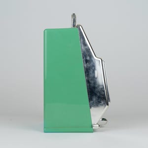 May include: A side view of a mint green kitchen appliance with a smooth, glossy finish. The front section features a polished, reflective chrome-colored metal panel with a stepped design. A small metal loop is visible at the top center of the unit.