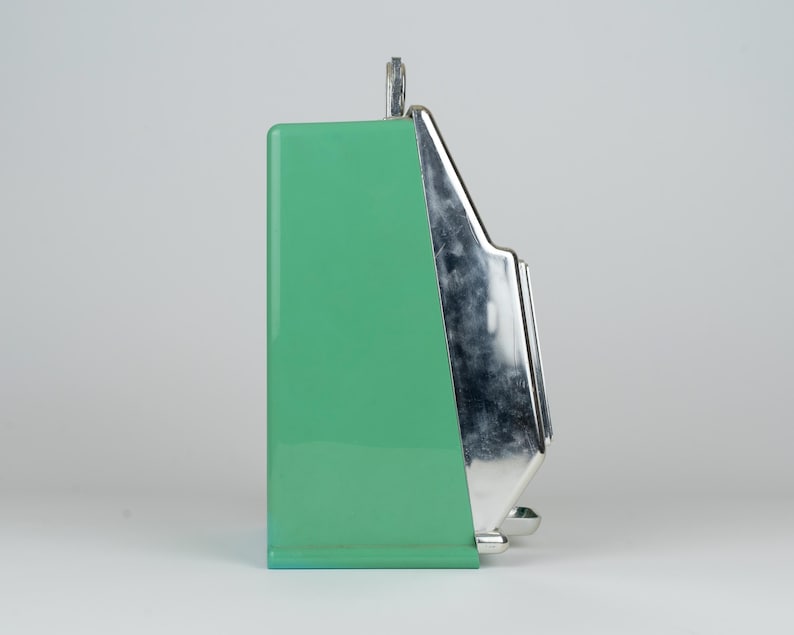 May include: A side view of a mint green kitchen appliance with a smooth, glossy finish. The front section features a polished, reflective chrome-colored metal panel with a stepped design. A small metal loop is visible at the top center of the unit.