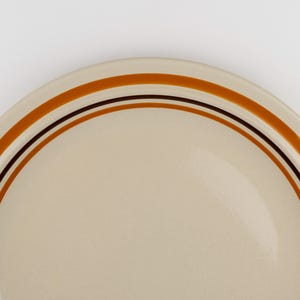 May include: A round ceramic plate with a smooth, light beige surface. The rim features a series of concentric circular bands in varying shades of brown and burnt orange, creating a simple, clean, and defined border around the edge of the plate.