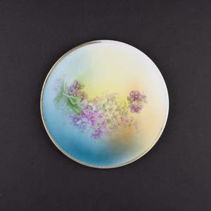 May include: A round porcelain plate featuring a hand-painted floral motif with clusters of purple and white flowers. The background displays a soft gradient transition from a deep blue on the left to a pale yellow on the right, finished with a thin silver rim.