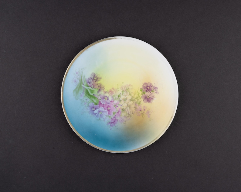 May include: A round porcelain plate featuring a hand-painted floral motif with clusters of purple and white flowers. The background displays a soft gradient transition from a deep blue on the left to a pale yellow on the right, finished with a thin silver rim.