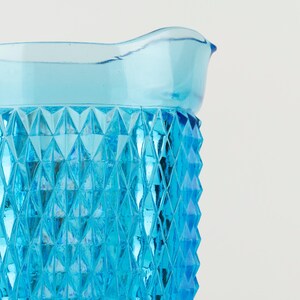 May include: A bright blue glass pitcher featuring a prominent diamond-shaped geometric pattern across its exterior surface. The vessel has a smooth, flared rim and a translucent finish that catches the light, highlighting the textured facets.
