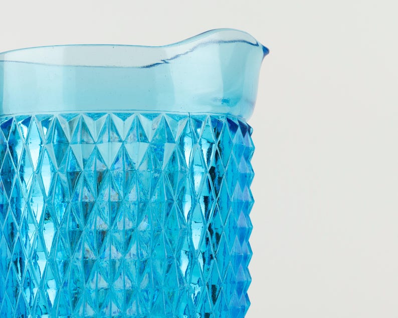 May include: A bright blue glass pitcher featuring a prominent diamond-shaped geometric pattern across its exterior surface. The vessel has a smooth, flared rim and a translucent finish that catches the light, highlighting the textured facets.